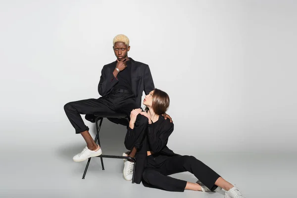 Trendy woman sitting on floor near blonde african american man on chair on grey with shadow — Stock Photo