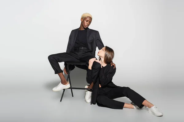 Trendy blonde woman on chair and brunette woman on floor looking at each other on grey with shadow — Stock Photo