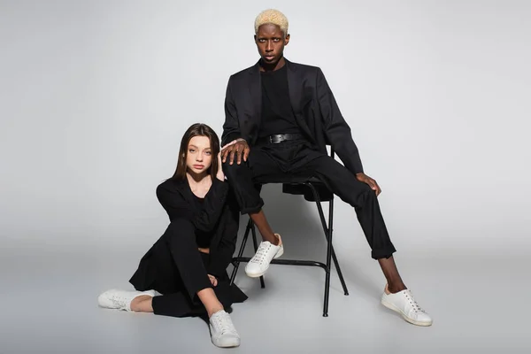 Blonde african american man sitting on chair near stylish woman on floor on grey with shadow — Stock Photo