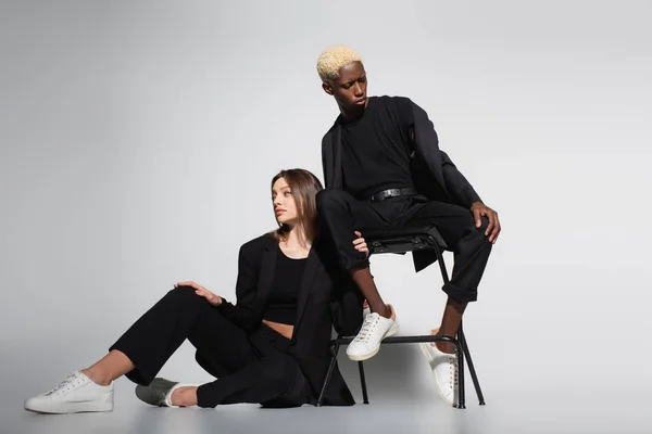 Mujer con estilo en el piso y mujer afroamericana rubia en silla posando en trajes negros en gris con sombra - foto de stock