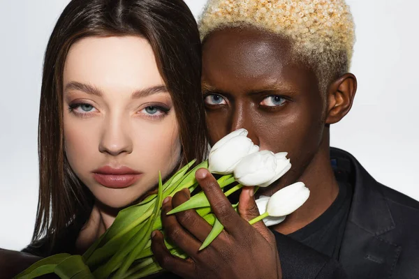 Close up view of young interracial couple looking at camera near white tulips isolated on grey — Stock Photo