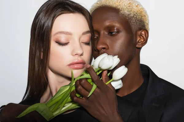 Blonde afrikanisch-amerikanische Mann hält weiße Tulpen neben hübsche Frau mit geschlossenen Augen isoliert auf grau — Stockfoto