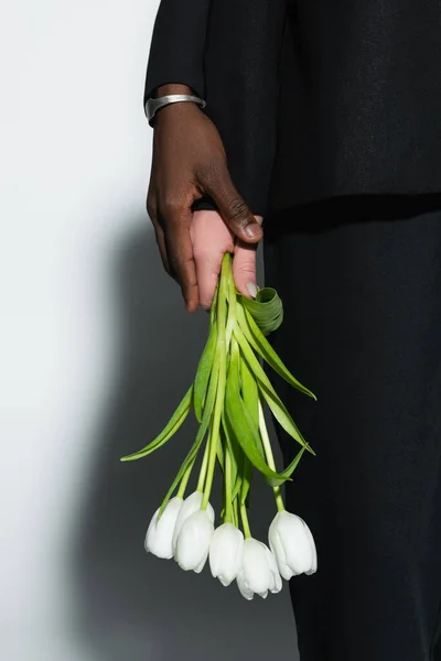 Обрезанный вид африканского амейкана, касающегося руки женщины, держащей белые тюльпаны на сером с тенью — стоковое фото