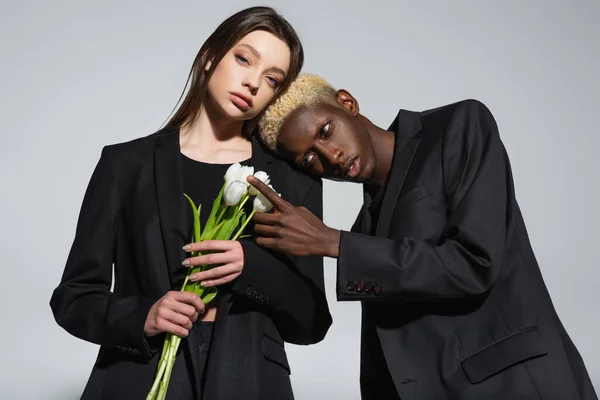 Homem americano africano elegante com cabelo branqueado tocando tulipas brancas perto de jovem mulher morena isolada em cinza — Fotografia de Stock