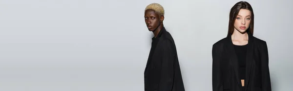 Jovem mulher em roupas pretas olhando para a câmera perto de homem americano africano com cabelo branqueado isolado em cinza, banner — Fotografia de Stock