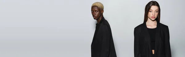 Mujer joven en ropa negra mirando a la cámara cerca de hombre afroamericano con el pelo blanqueado aislado en gris, pancarta — Stock Photo