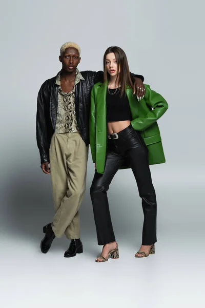 Visão de comprimento total de casal interracial elegante em jaquetas de couro olhando para a câmera no cinza — Fotografia de Stock