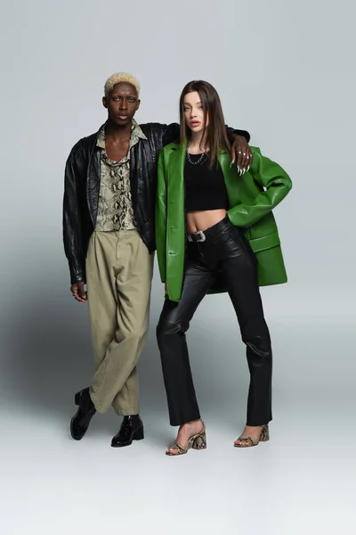 Full length view of stylish interracial couple in leather jackets looking at camera on grey — Stock Photo