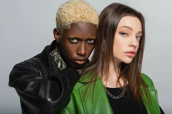 Brunette woman and blonde african american man in leather jackets looking at camera isolated on grey — Stock Photo