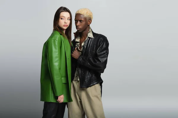 Blonde african american man and brunette woman in leather jackets standing on grey — Stock Photo