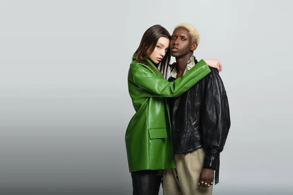 Bonita mujer en chaqueta de cuero verde abrazando elegante hombre afroamericano en gris - foto de stock