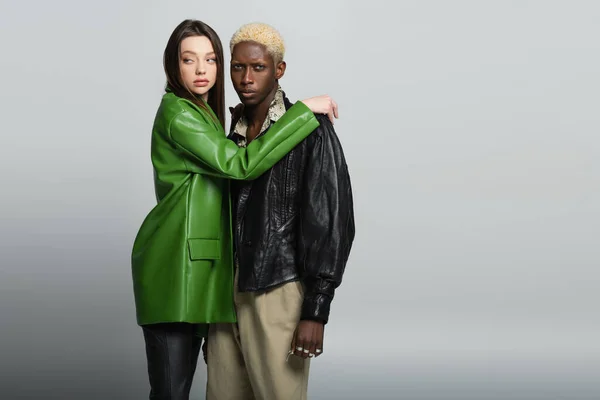 Mujer con estilo en chaqueta de cuero verde abrazando hombre afroamericano rubia mirando a la cámara en gris - foto de stock