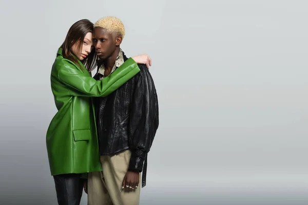 Young woman in green leather jacket hugging blonde african american man and looking at camera on grey — Stock Photo