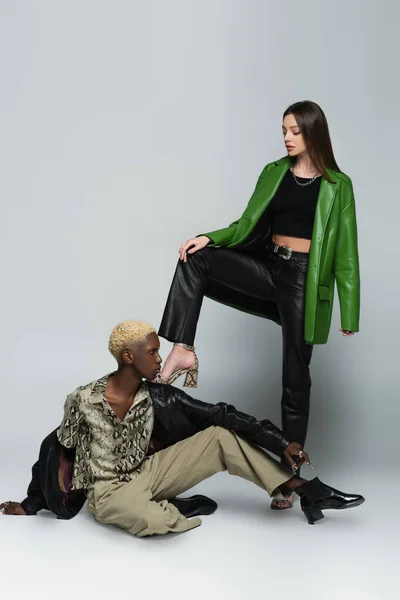 Stylish brunette woman stepping on blonde african american man sitting on grey — Stock Photo