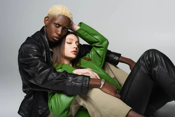 Stylish african american man embracing sensual brunette woman while sitting on grey — Stock Photo