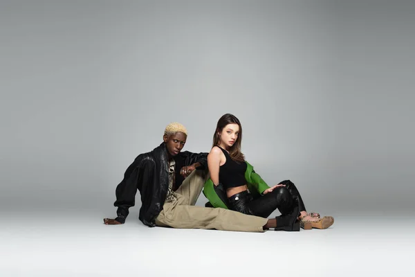 Sensual woman in black top and leather trousers sitting near blonde african american man on grey — Stock Photo