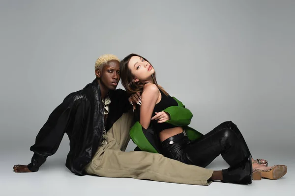 Multiethnic couple in fashionable clothes looking at camera while sitting on grey — Stock Photo