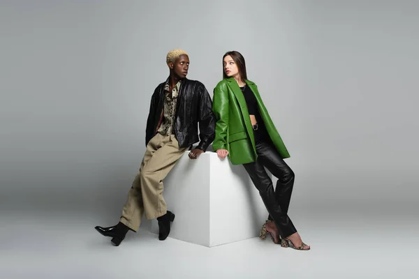 Full length view of fashionable interracial couple sitting on white cube and looking at each other on grey — Stock Photo