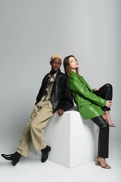 Young brunette woman and blonde african american man sitting on white cube and looking at camera on grey — Stock Photo