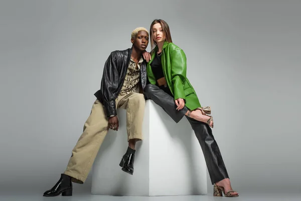 Brunette woman and blonde african american man in leather jackets sitting on white cube on grey — Stock Photo