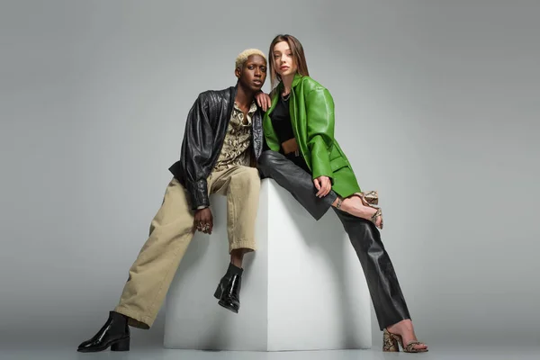 Morena mujer y rubia afroamericana hombre en chaquetas de cuero sentado en cubo blanco en gris - foto de stock