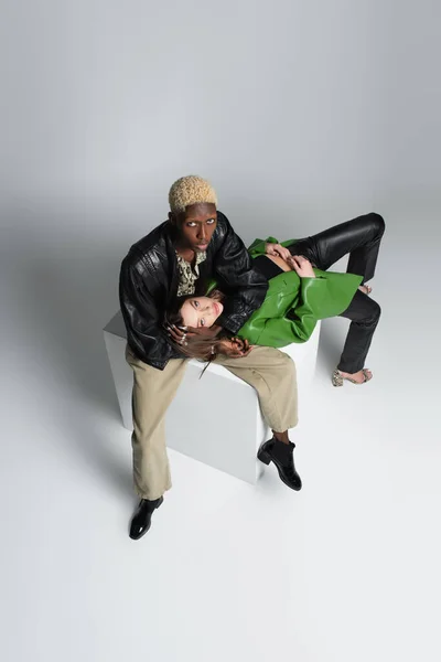 Overhead view of brunette woman and blonde african american man sitting on white cube on grey — Stock Photo
