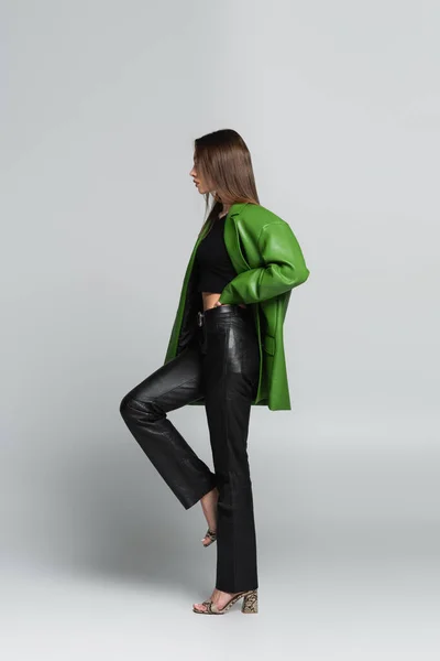 Vue latérale de la femme en sandales, pantalon et veste en cuir debout sur une jambe avec la main à la taille sur gris — Photo de stock