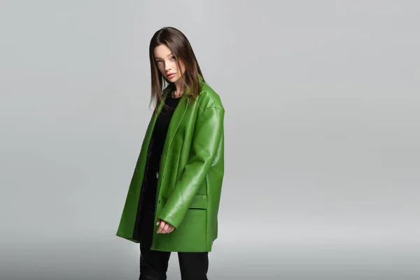 Молода і стильна жінка в зеленій шкіряній куртці дивиться на камеру ізольовано на сірому — стокове фото