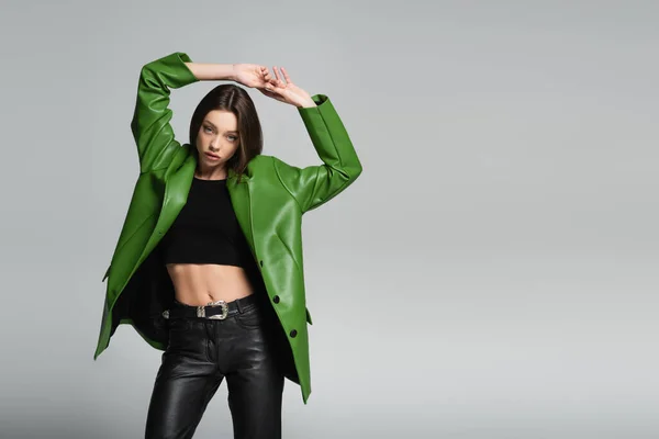 Trendy woman in black top and green leather jacket standing with hands above head isolated on grey — Stock Photo