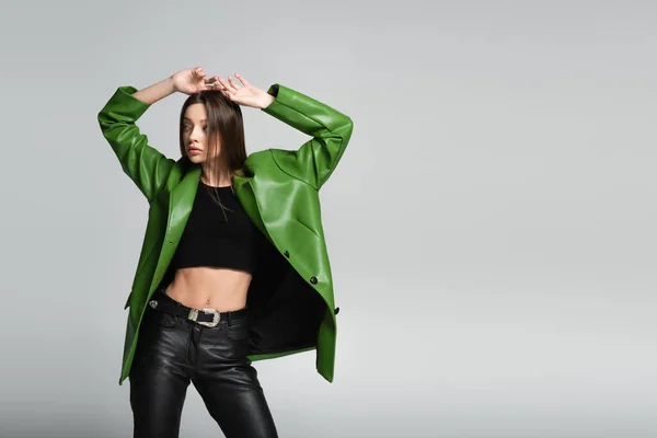 Mujer de moda en top negro, pantalones de cuero y chaqueta posando con las manos sobre la cabeza aisladas en gris - foto de stock