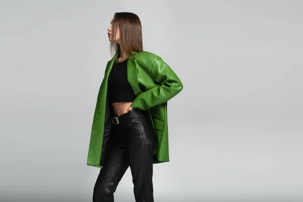 Mujer morena en pantalones de cuero negro y chaqueta verde posando con la mano en la cintura aislada en gris - foto de stock