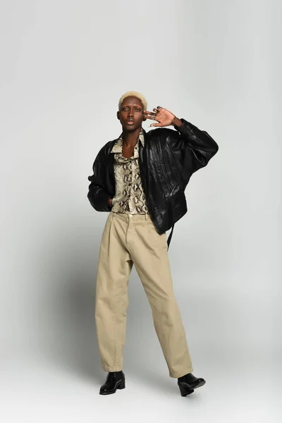 Vista completa del hombre afroamericano rubio en chaqueta negra y pantalones beige mirando a la cámara en gris - foto de stock