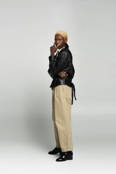 Vista integrale dell'uomo biondo afroamericano in giacca di pelle nera e pantaloni beige su grigio — Foto stock