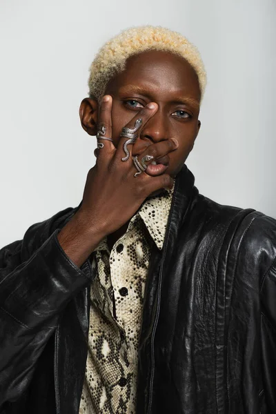 Stylish african american man in snake-shaped rings posing with hand on face isolated on grey — Stock Photo