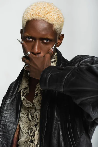 Blonde and stylish african american man with metal fingernails covering mouth with hand isolated on grey — Stock Photo