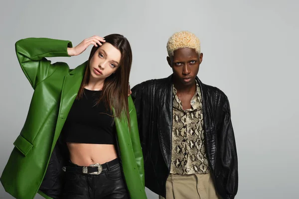 Young woman in black top and green leather jacket posing near blonde african american man isolated on grey — Stock Photo