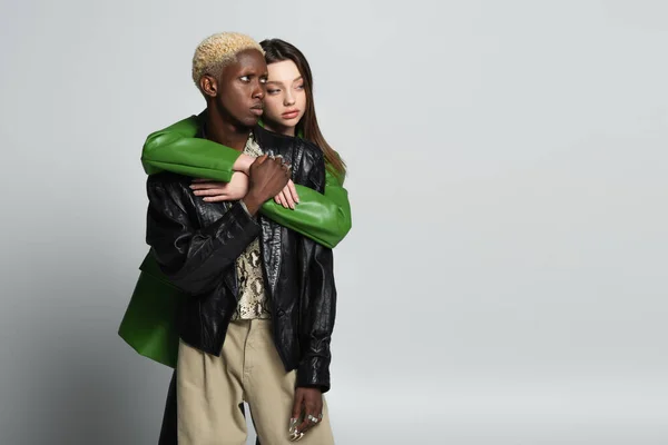 Brunette woman embracing blonde african american man in trendy clothes and looking away on grey — Stock Photo