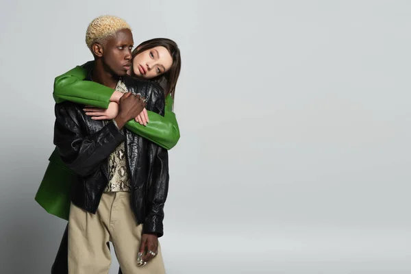 Young woman looking at camera while hugging stylish african american man with bleached hair on grey — Stock Photo
