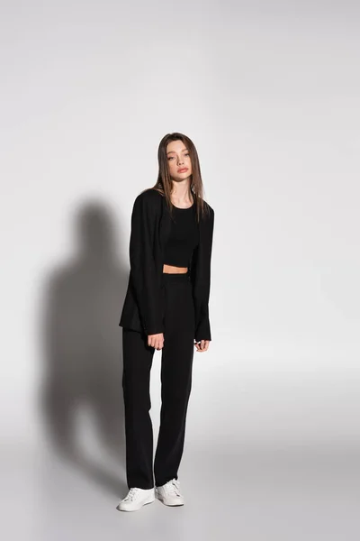 Full length view of brunette woman in black suit and white sneakers on grey with shadow — Stock Photo