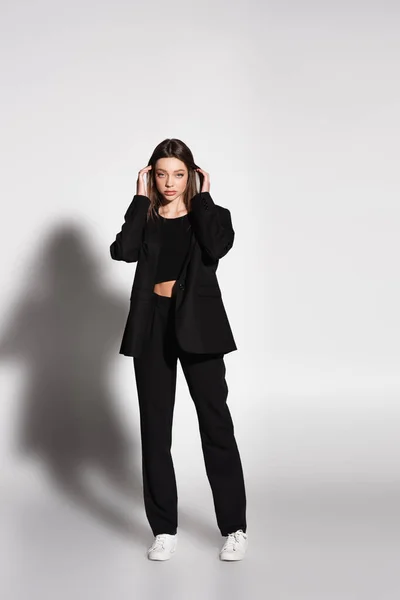Ganzkörperansicht einer Frau im schwarzen Anzug und weißen Turnschuhen, die ihr Haar auf grau mit Schatten fixiert — Stockfoto