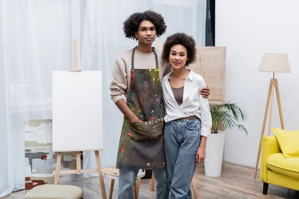 Joven pareja afroamericana sonriendo a la cámara cerca del caballete en casa — Stock Photo