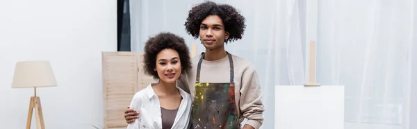 Joven afroamericano hombre abrazando novia cerca caballete en casa, pancarta — Stock Photo