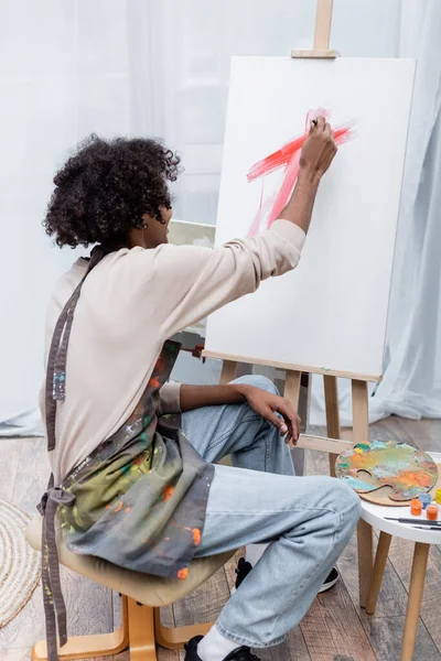 Joven afroamericano hombre en delantal pintura sobre lienzo en casa - foto de stock