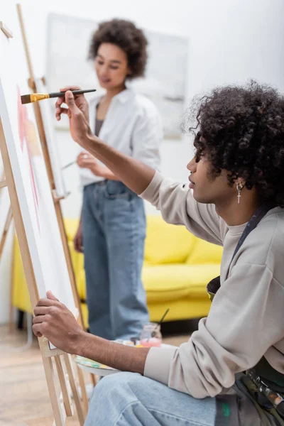Jovem afro-americano em avental pintura sobre tela perto de namorada borrada em casa — Fotografia de Stock