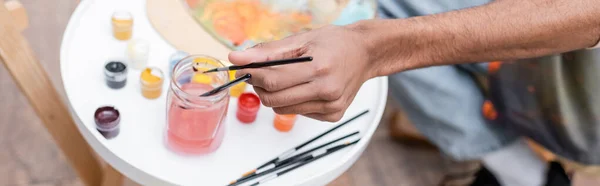 Top view of african american man holding paintbrush near water and paints at home, banner — Stock Photo