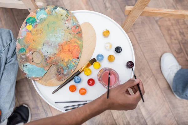 Top view of african american man holding paintbrush near paints and palettes at home — Stock Photo