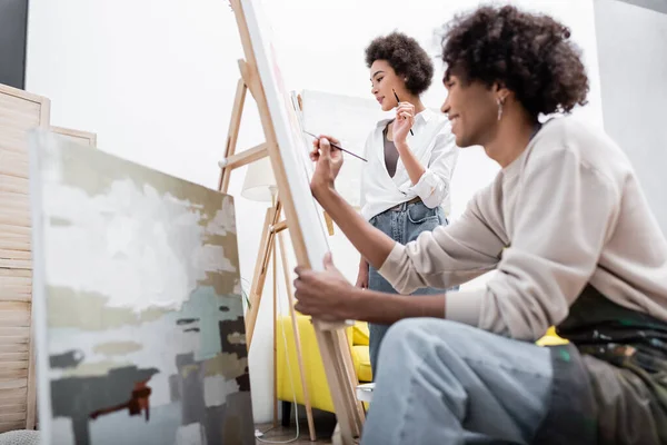 Jovem afro-americana segurando pincel perto de tela e namorado borrado pintando em casa — Fotografia de Stock