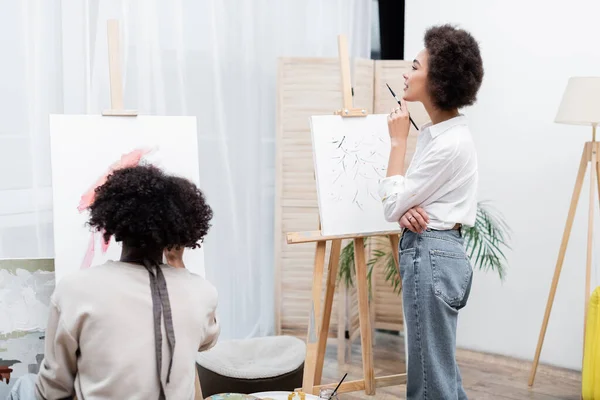 Offensiv afrikanisch-amerikanische Frau hält Pinsel in der Nähe von Leinwand und Freund zu Hause — Stockfoto