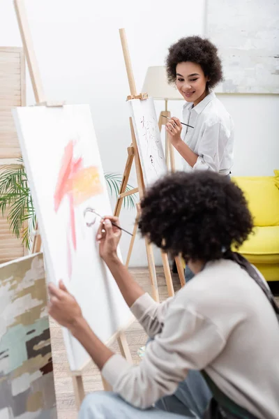 Sonriente mujer afroamericana pintura sobre lienzo cerca borrosa novio en casa - foto de stock