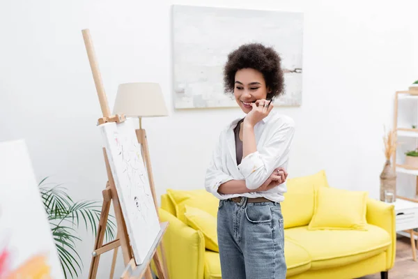 Sonriente mujer afroamericana mirando lienzo en caballete en casa - foto de stock